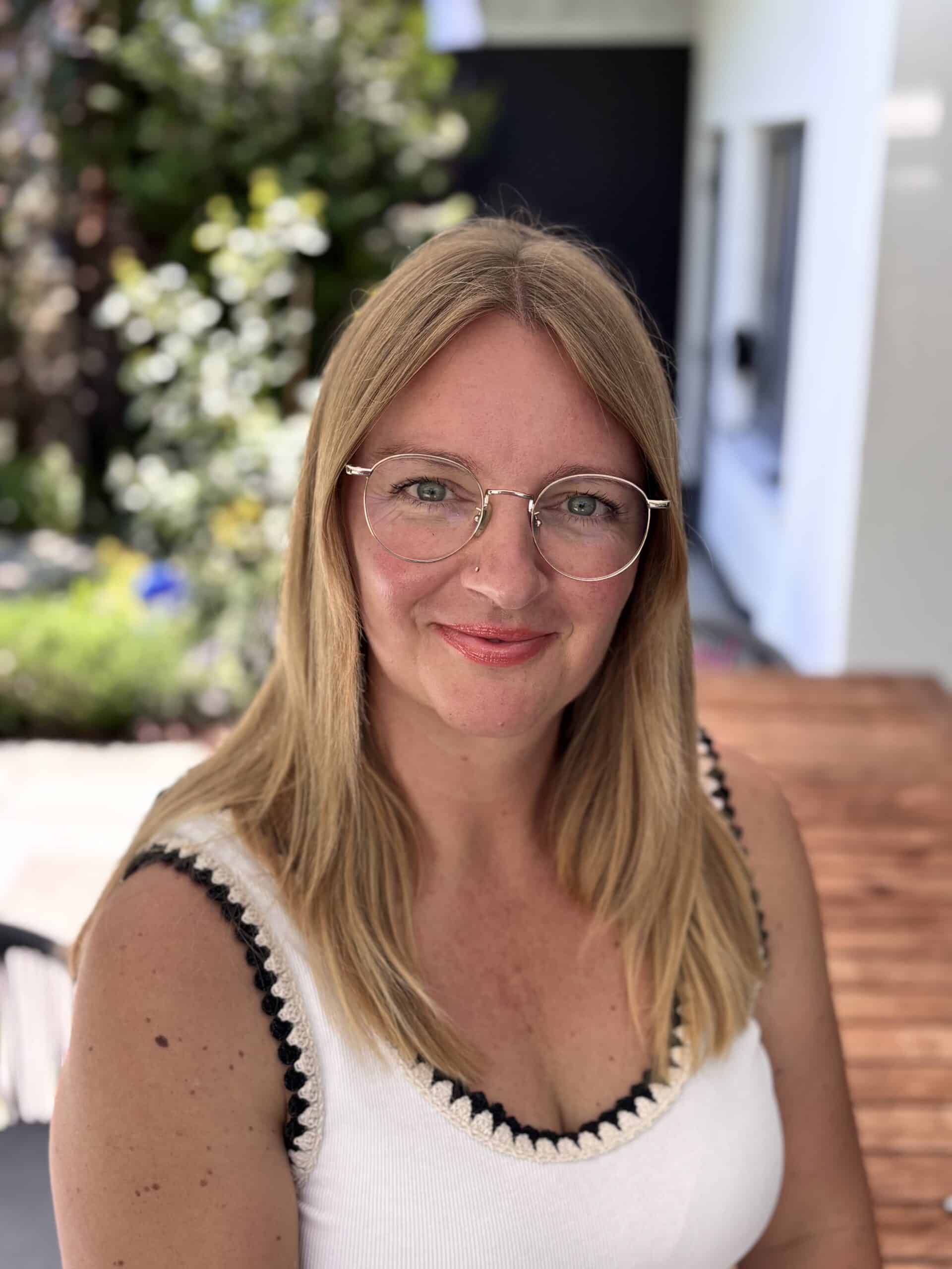 Alexandra Reiter mit Brille im Garten, lächelnd, natürliche Umgebung.